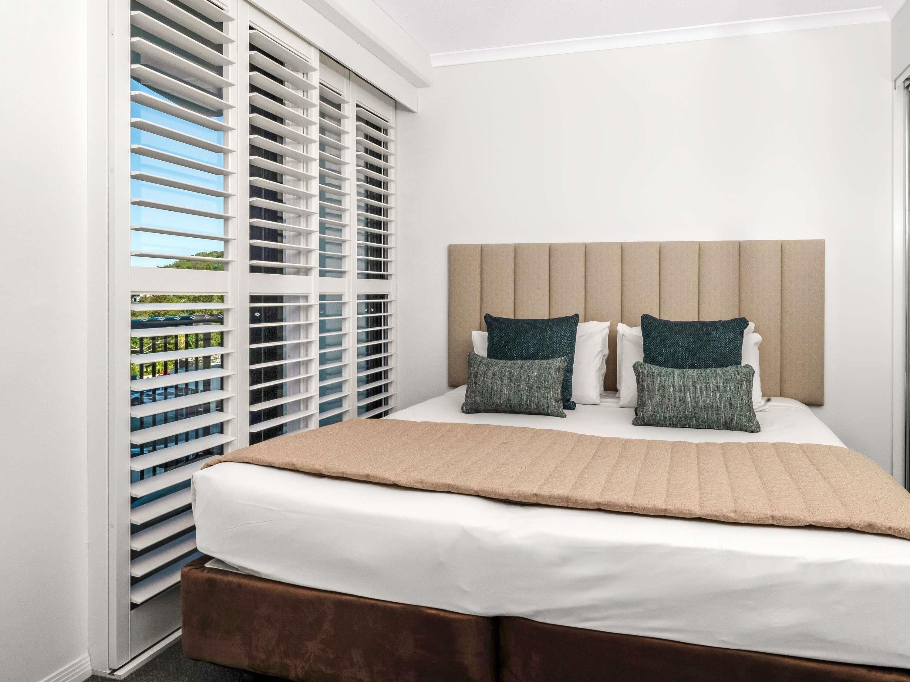 A bedroom with a bed, two pillows, a headboard, and a window with blinds.