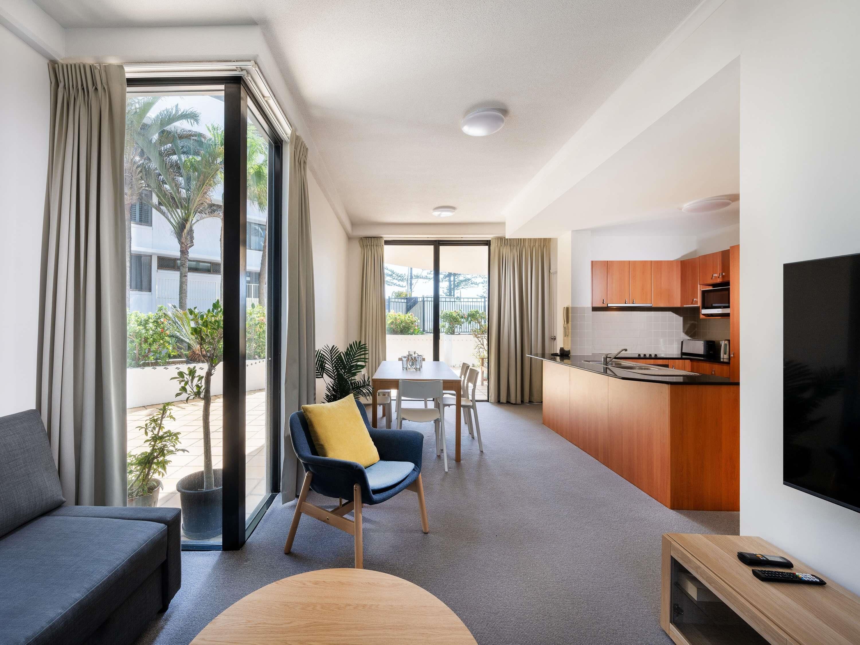 A modern living room with a sofa, a dining area with a table and chairs, a kitchen with wooden cabinets and a TV, and large windows with curtains.