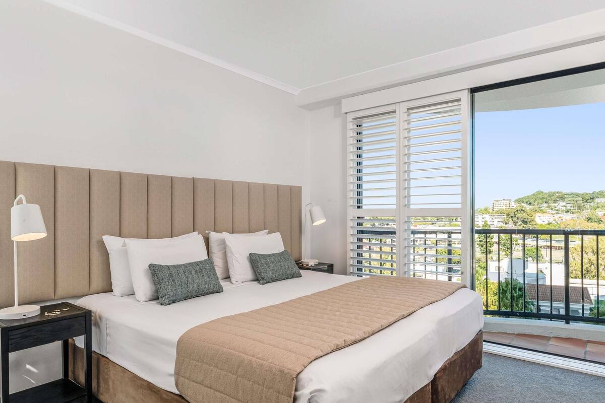 A modern hotel room with a large bed, two bedside tables, a nightstand, and a balcony with a view.