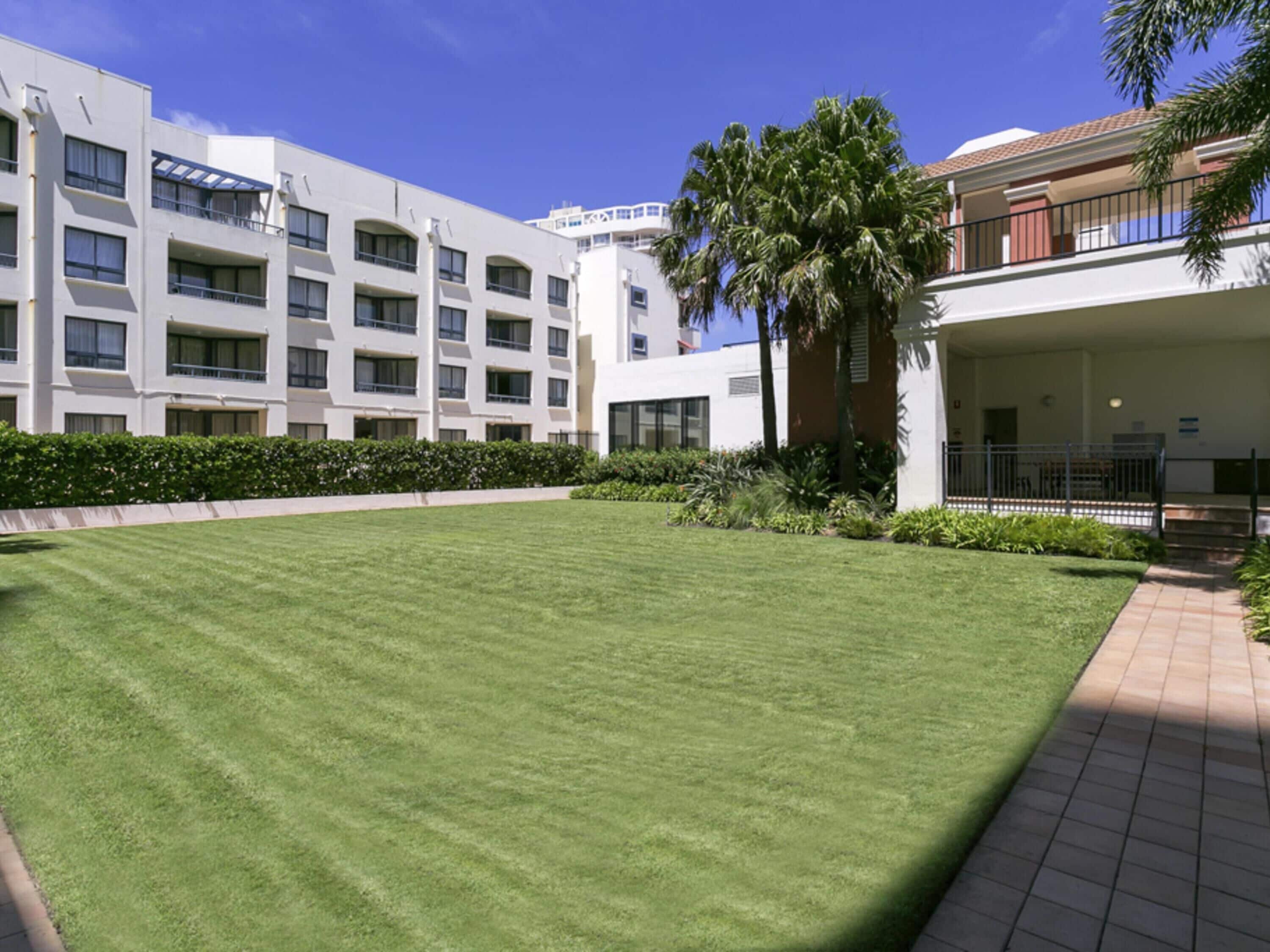 A well-maintained courtyard with a lawn, surrounded by modern buildings with balconies and a clear blue sky.