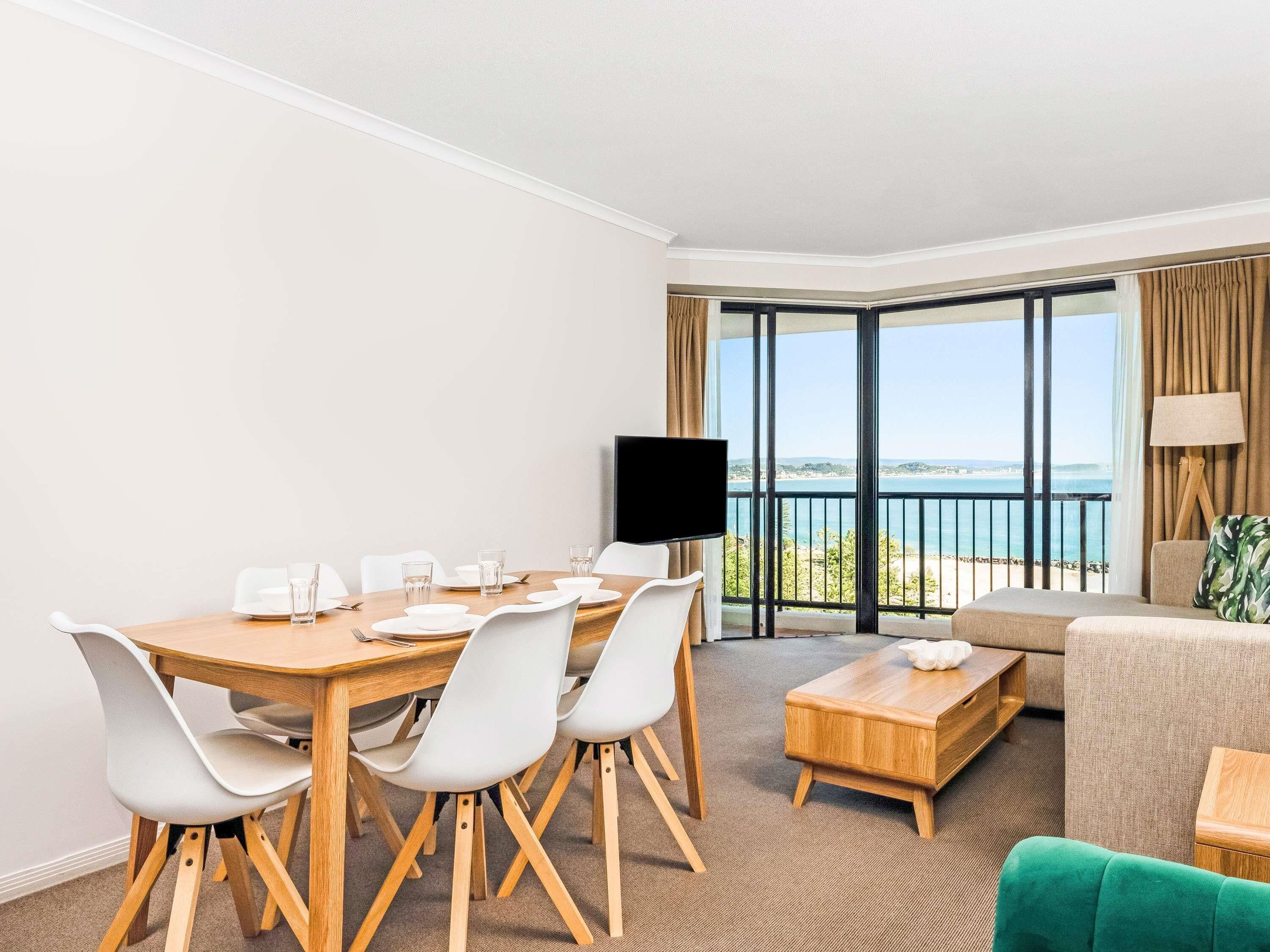 A hotel room with a dining table set for four, a sofa, a TV, and a balcony with a view.