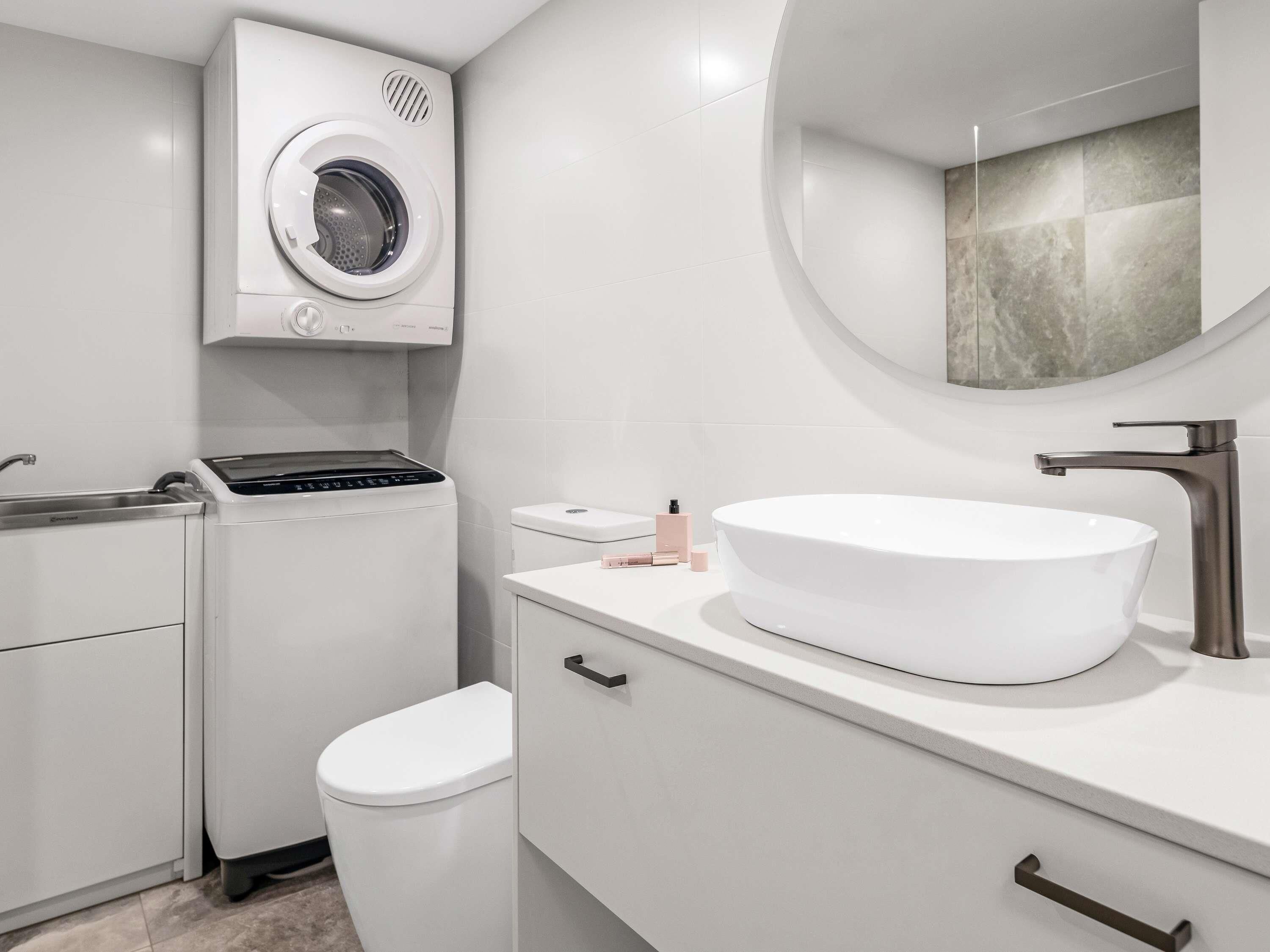 A modern bathroom with a built-in washing machine, a toilet, a sink with a round mirror, and a countertop with toiletries.