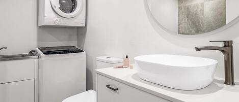 A modern bathroom with a built-in washing machine, a toilet, a sink with a round mirror, and a countertop with toiletries.