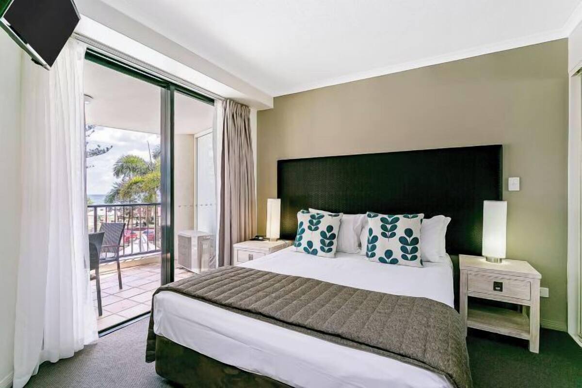 A hotel room with a bed, a nightstand, a TV, a balcony with chairs, and a view of the outdoors.