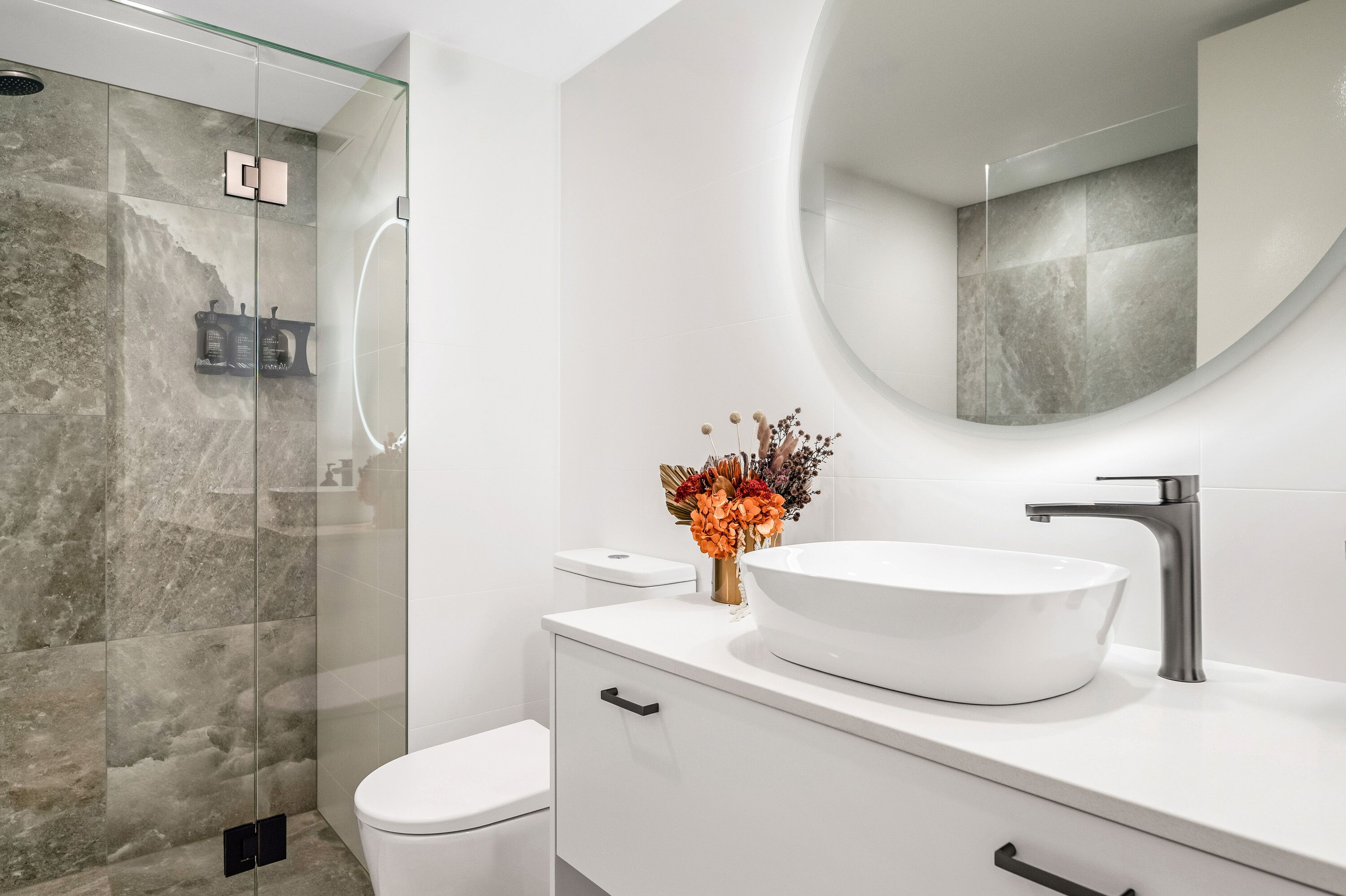 Une salle de bain moderne avec une douche à parois de verre, un lavabo blanc et un miroir rond.