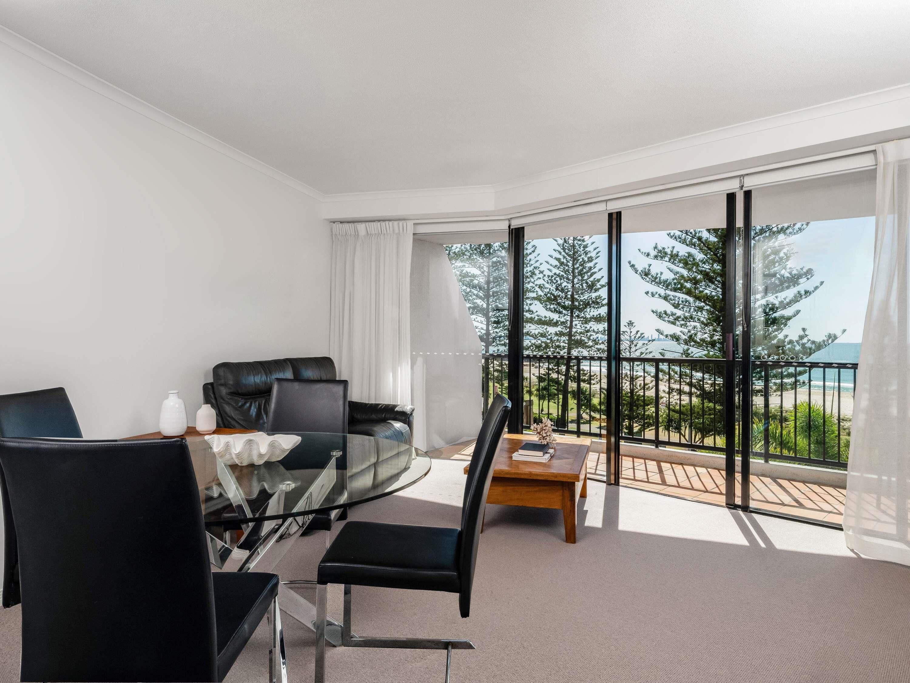Una habitación de hotel moderna con balcón, una mesa de comedor de cristal, sillas negras, un sofá de cuero y vistas al mar y árboles.