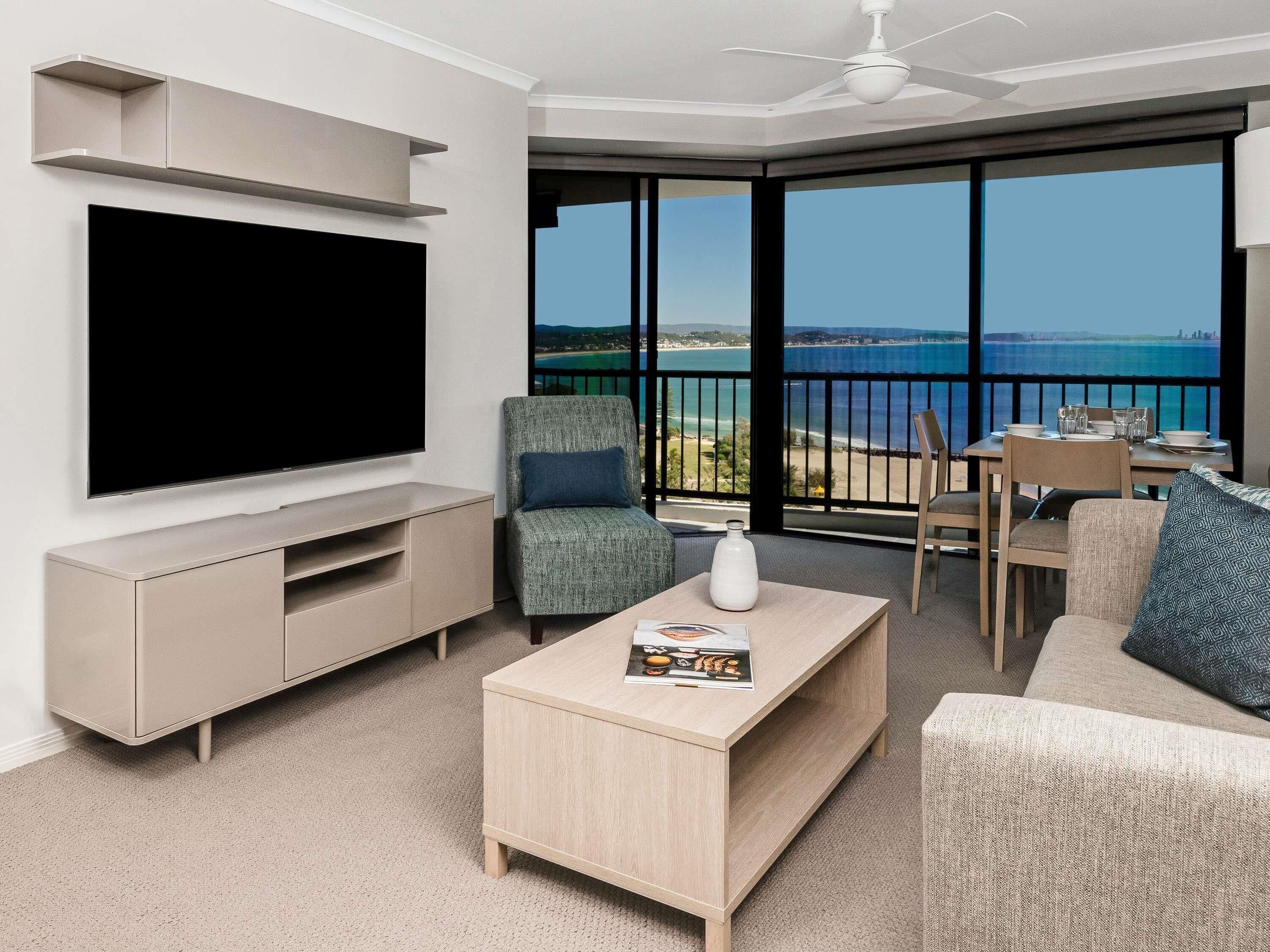 A modern living room with a flat-screen TV, a coffee table, a sofa, and a dining area with a view of the ocean.