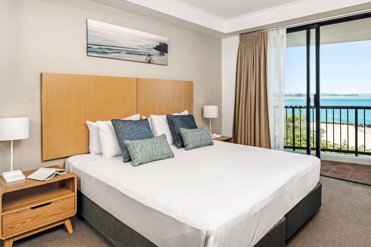 A hotel room with a large bed, a nightstand, a lamp, a painting, and a balcony with a view of the sea.