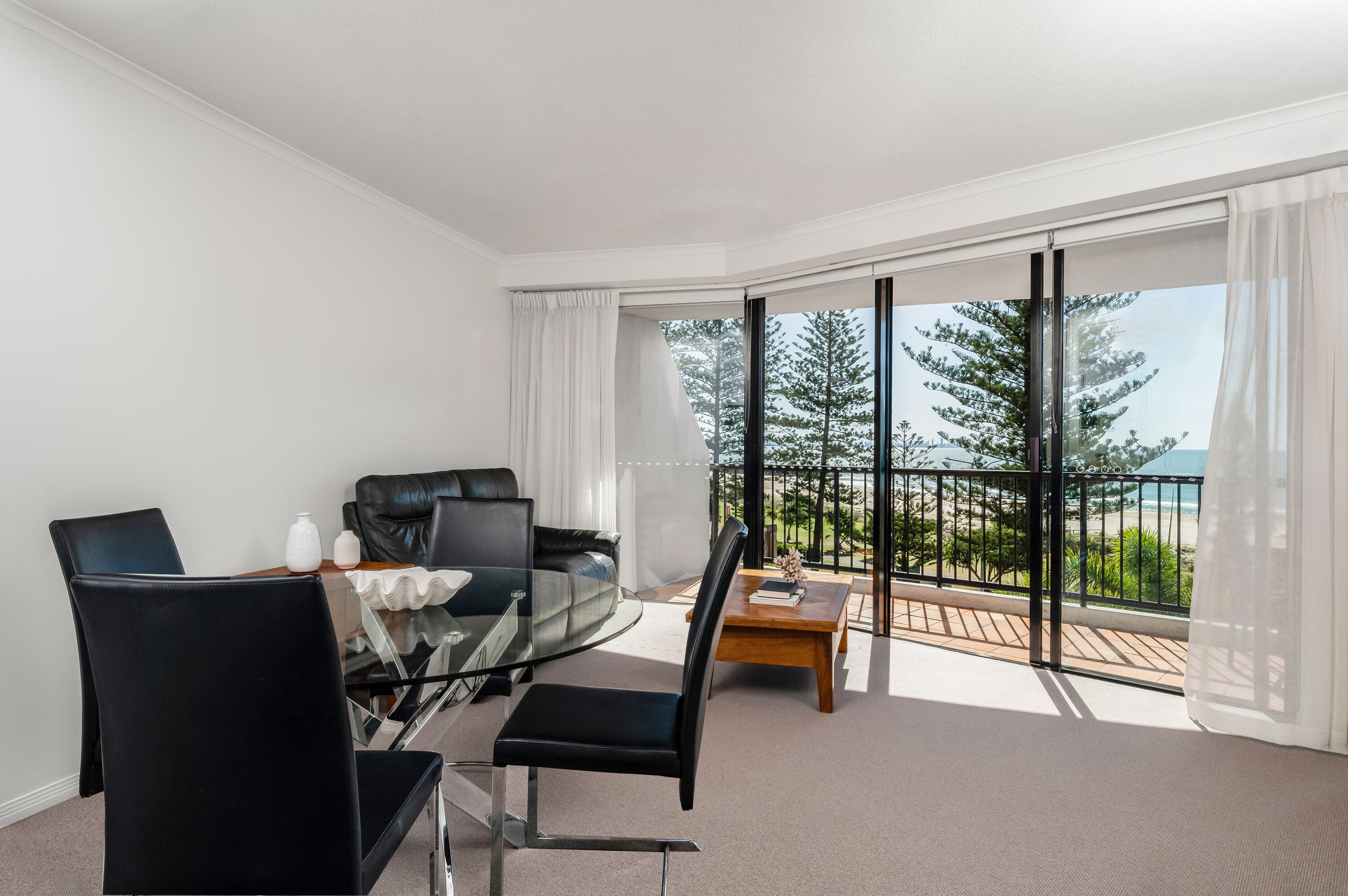 Una habitación moderna con mesa de vidrio, sillas negras, sofá de cuero y un amplio ventanal con vistas a árboles y una playa.