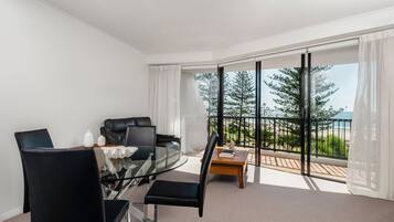 Una habitación moderna con mesa de vidrio, sillas negras, sofá de cuero y un amplio ventanal con vistas a árboles y una playa.