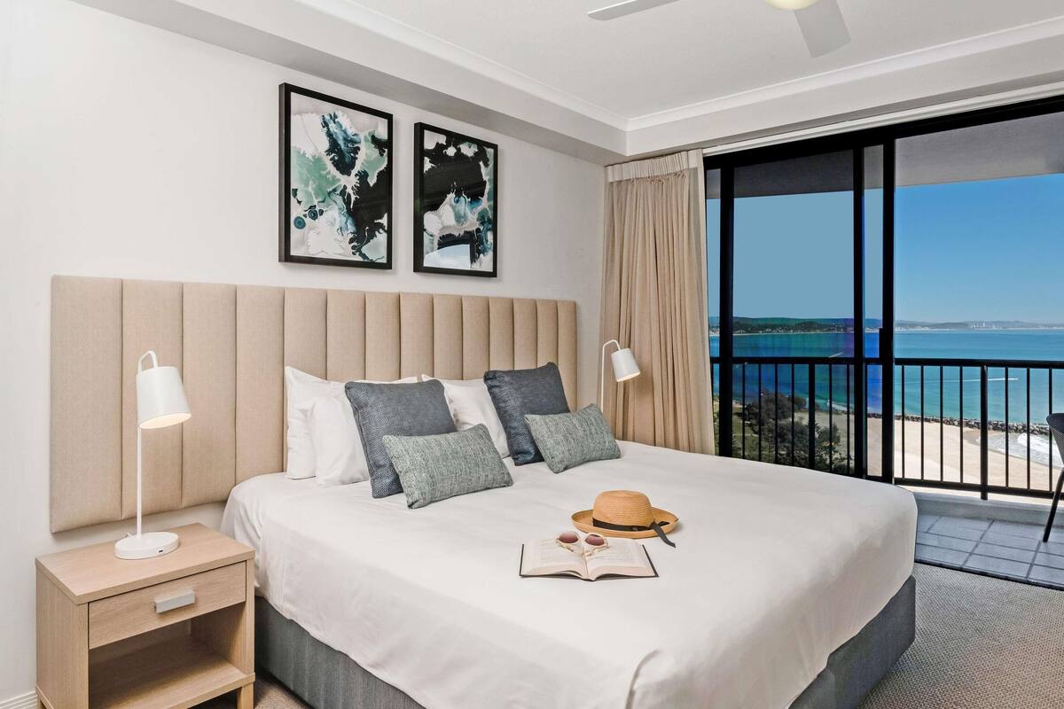 A hotel room with a bed, nightstands, a ceiling fan, artwork, a view of the sea, and a balcony.