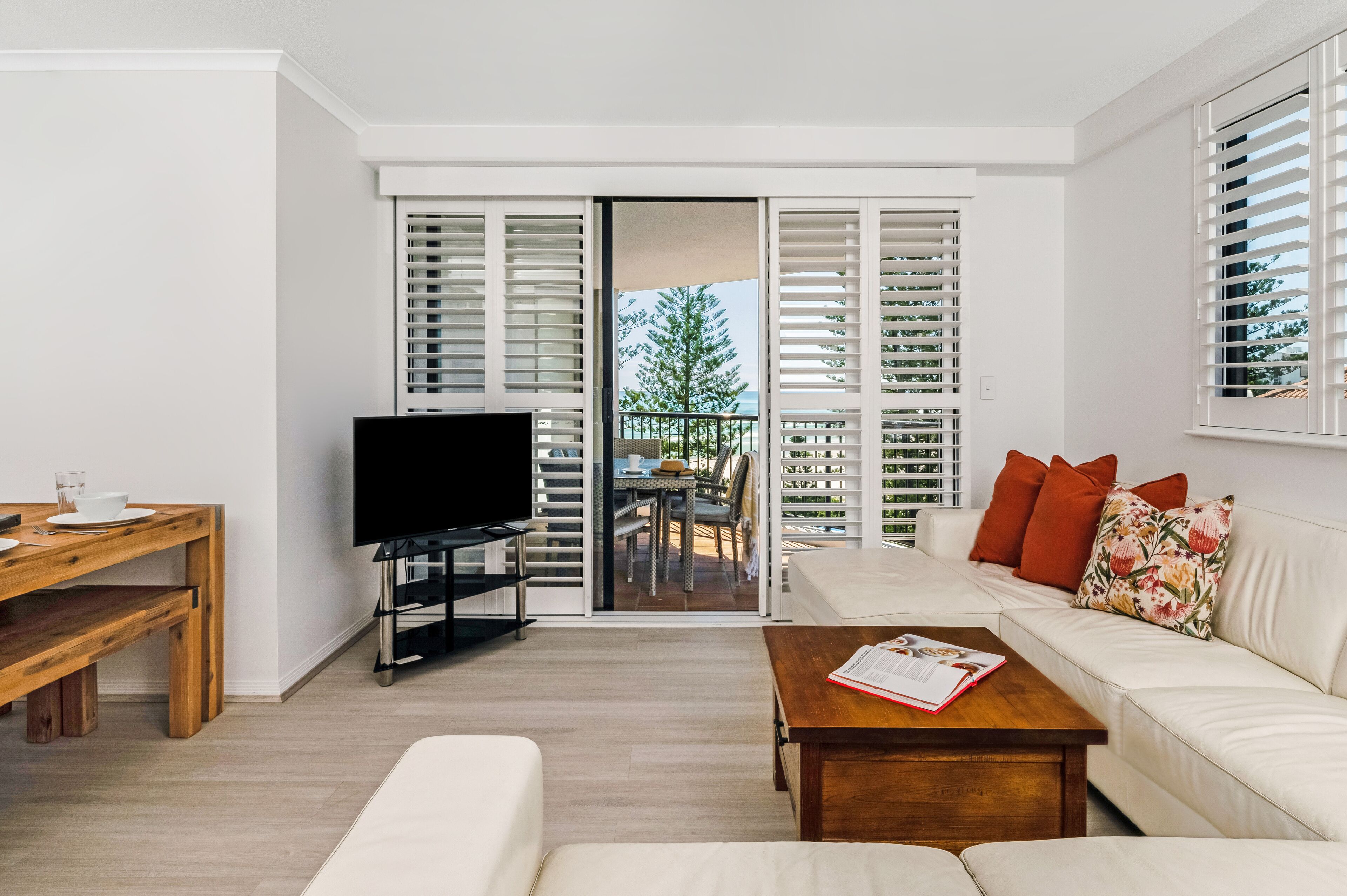 A modern living room with a flat-screen TV, a wooden coffee table, a white sofa, and a balcony with white shutters.