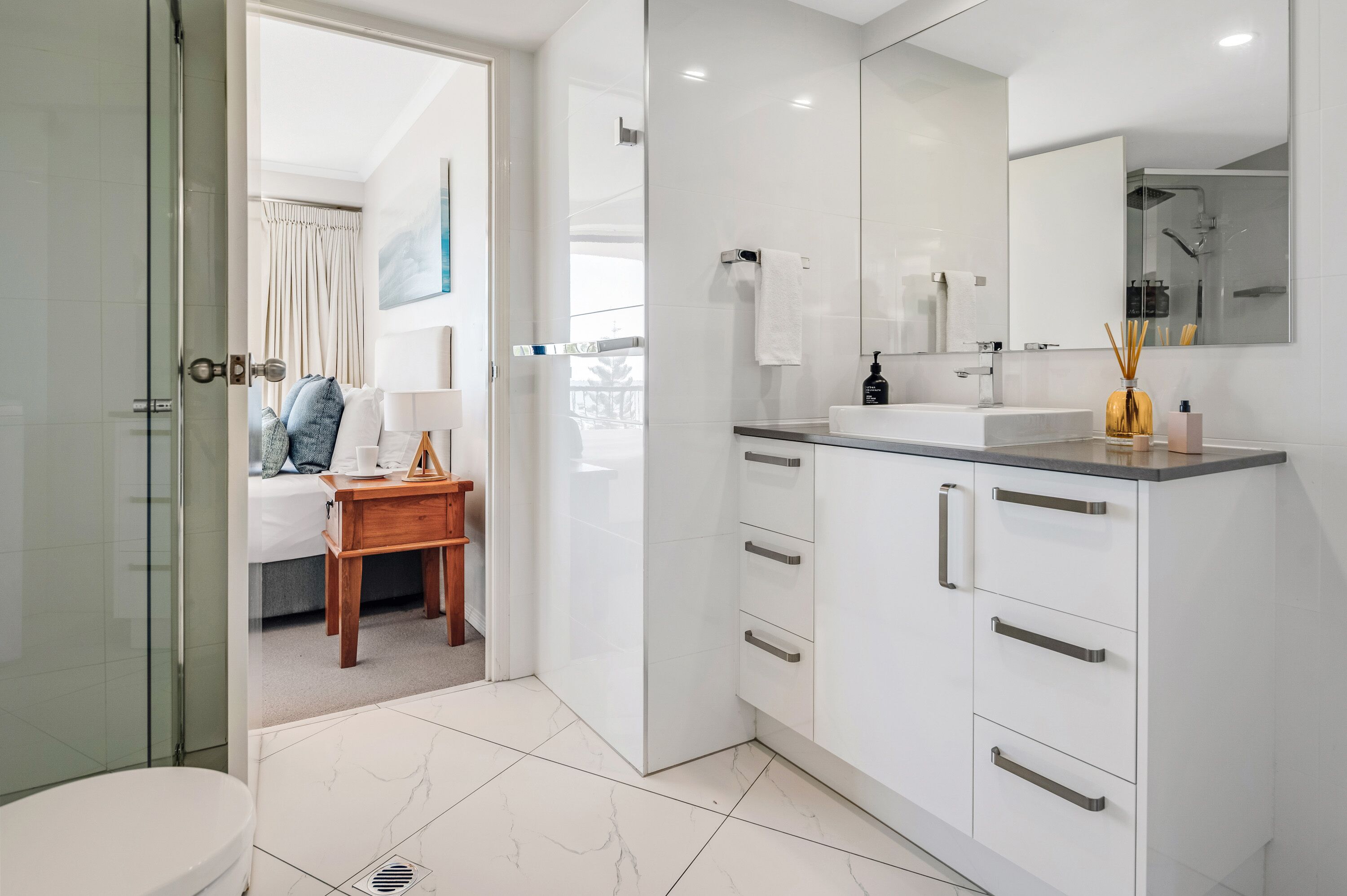 A modern bathroom with a glass shower enclosure, a white sink with a mirror, and a wooden bedside table with a lamp.