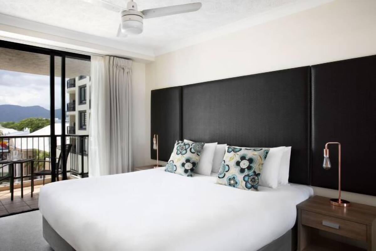 Ein Hotelzimmer mit einem großen Bett, einem Deckenventilator, einem Balkon mit Blick auf Gebäude und Berge sowie einem Nachttisch mit einer Lampe.