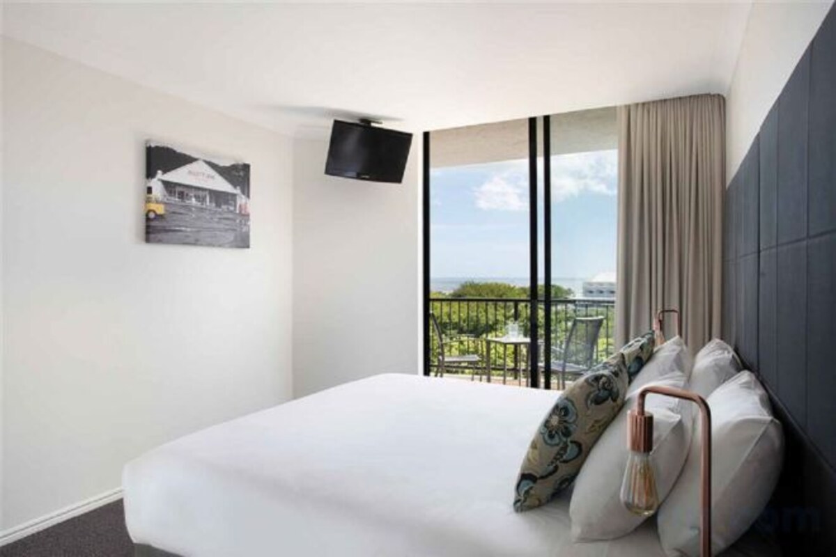 A hotel room with a bed, a TV mounted on the wall, a balcony with a view, and a table with chairs outside.