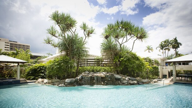 A swimming pool with a waterfall feature surrounded by tropical vegetation and palm trees. - Mantra Esplanade (Cairns)