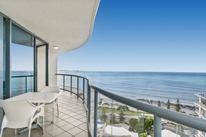 A balcony with a white table and chairs, overlooking a coastal view with the sea and a rocky shoreline.