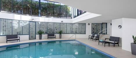 Piscina interior con vistas a árboles a través de amplias ventanas, rodeada de zonas de estar y plantas en macetas.