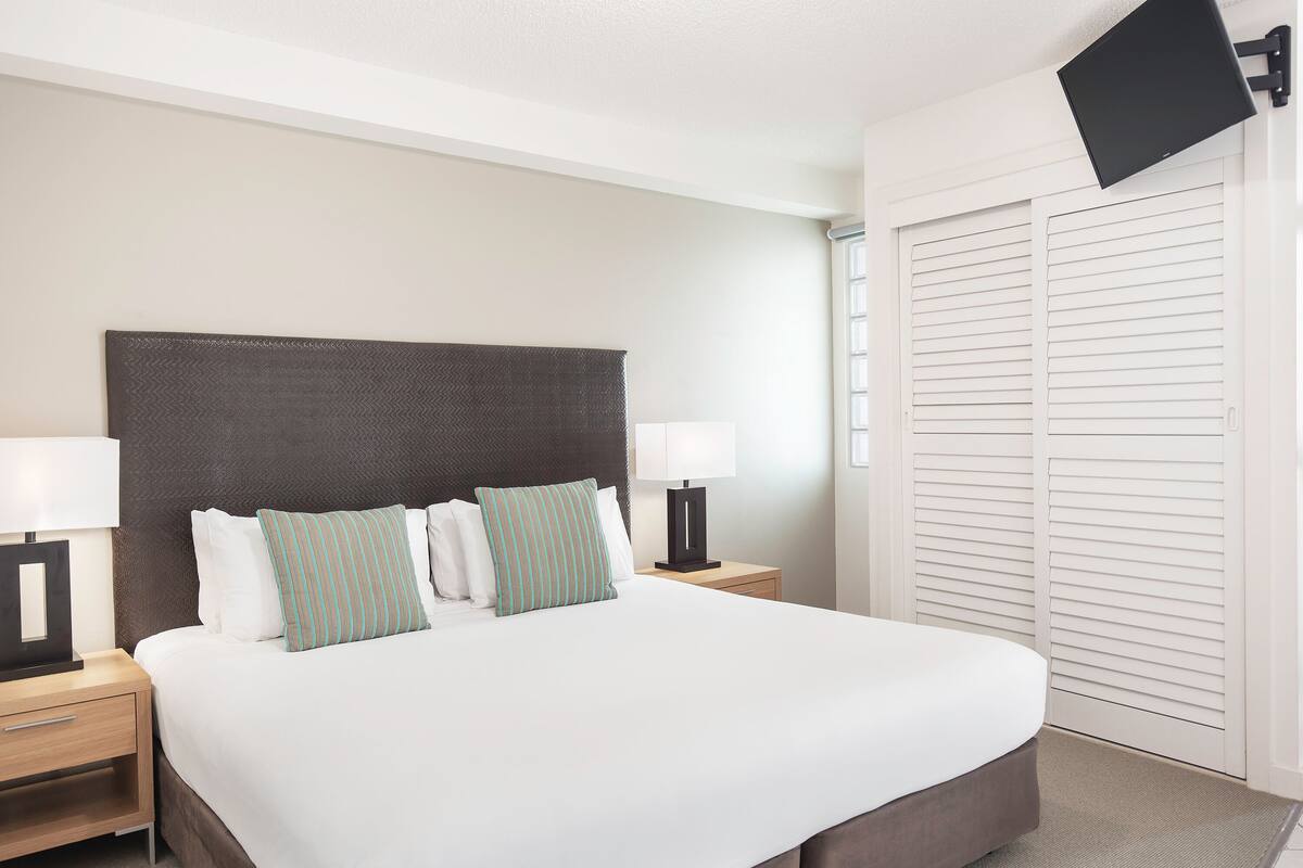 Una cama bien hecha con ropa de cama blanca y almohadas a rayas, flanqueada por mesitas de noche con lámparas, en una habitación con cabecera oscura y paredes blancas.