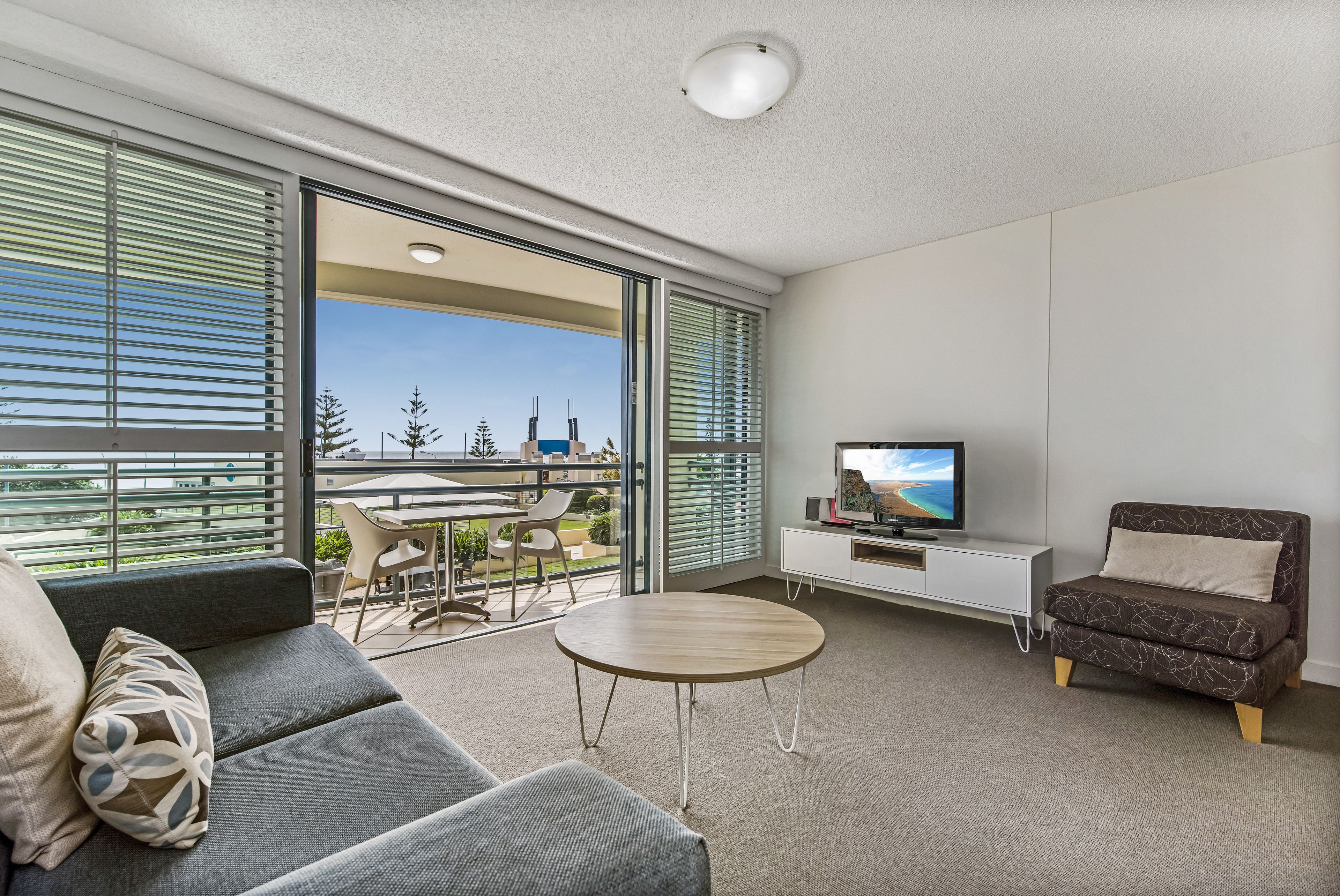 A modern living room with a sofa, a round coffee table, a TV, and a balcony with outdoor furniture.