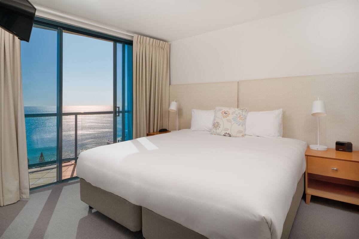 Habitación de hotel con una cama grande, dos mesitas de noche, vistas al océano y un balcón.