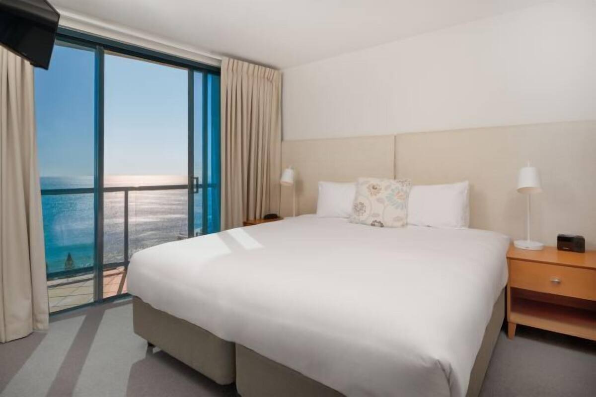 Una habitación de hotel con una cama grande, una mesita de noche, una lámpara y vista al océano.