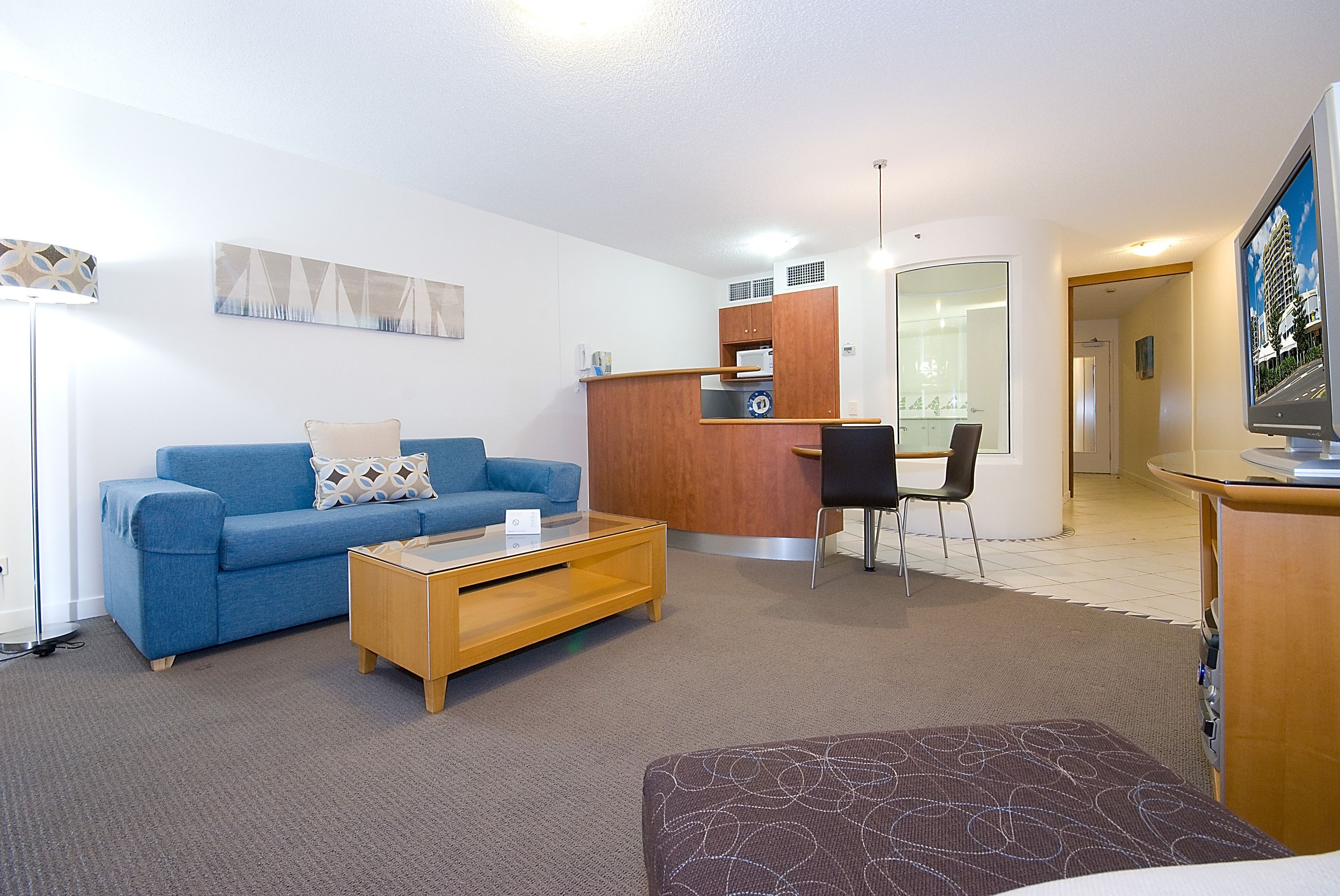 A modern living room with a blue sofa, a wooden coffee table, and a brown ottoman. There is a small kitchen area with a refrigerator, a microwave, and a sink.