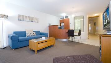 A modern living room with a blue sofa, a wooden coffee table, and a brown ottoman. There is a small kitchen area with a refrigerator, a microwave, and a sink.