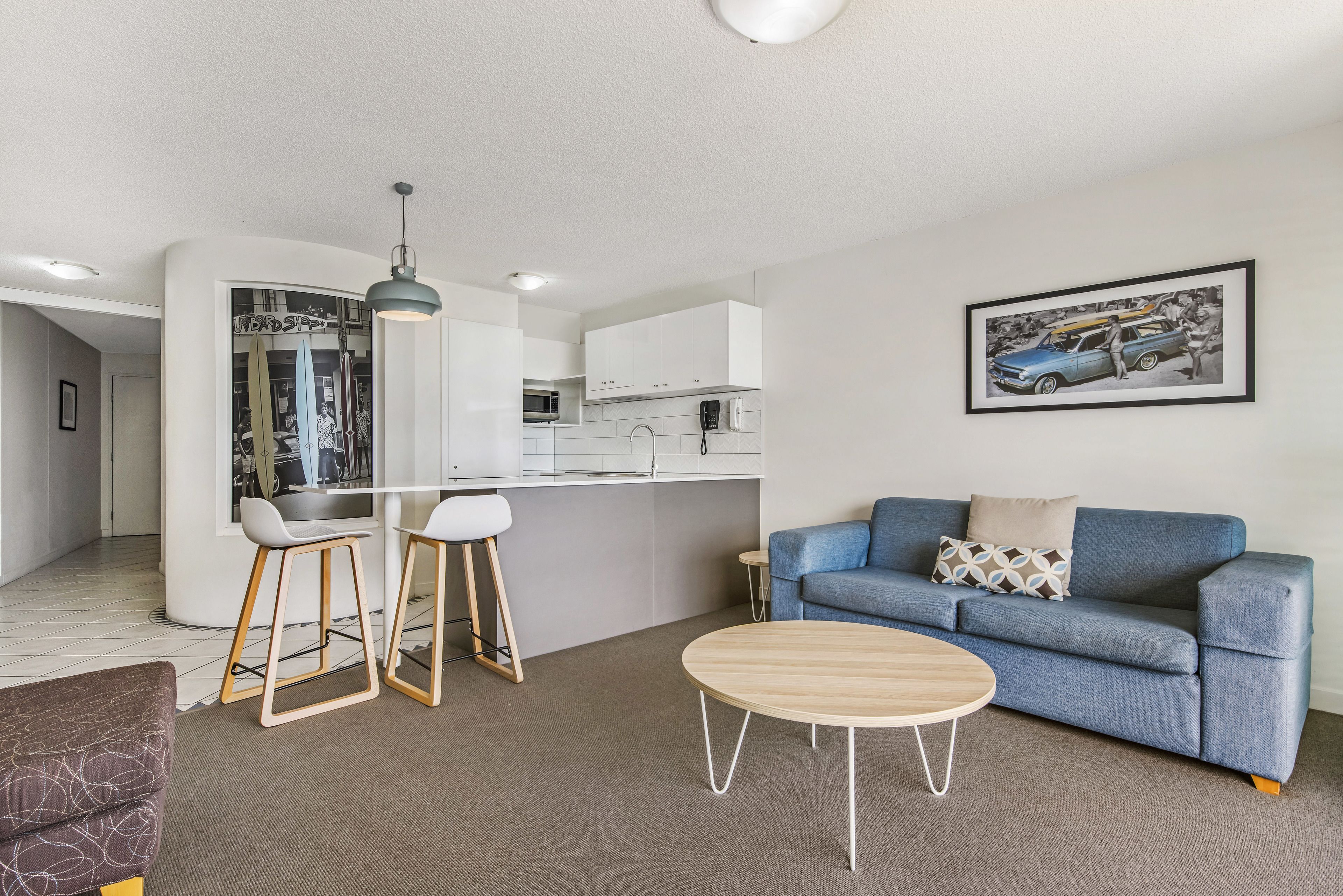A modern living room with a blue sofa, a wooden coffee table, and a kitchen area with white cabinets and a bar stool.