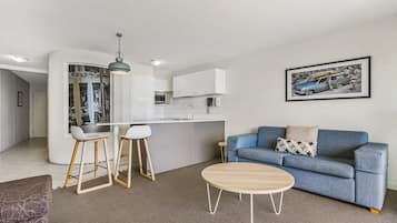 Una sala de estar moderna con un sofá azul, una mesa de centro de madera y una cocina con armarios blancos y un taburete de bar.