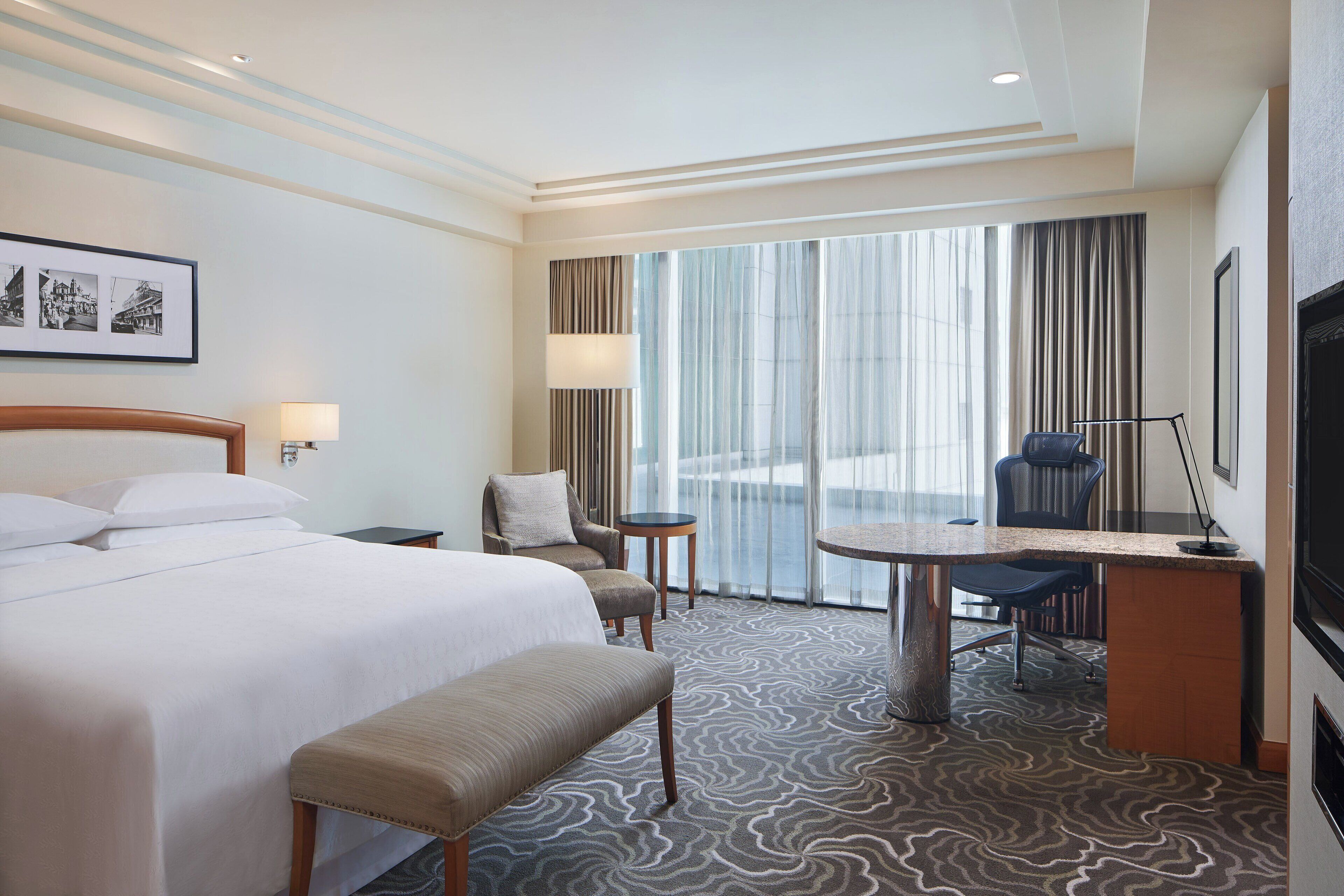 Ein Hotelzimmer mit einem großen Bett, einem Schreibtisch mit Stuhl, einer Bank, einem kleinen Tisch und einem Fernseher.