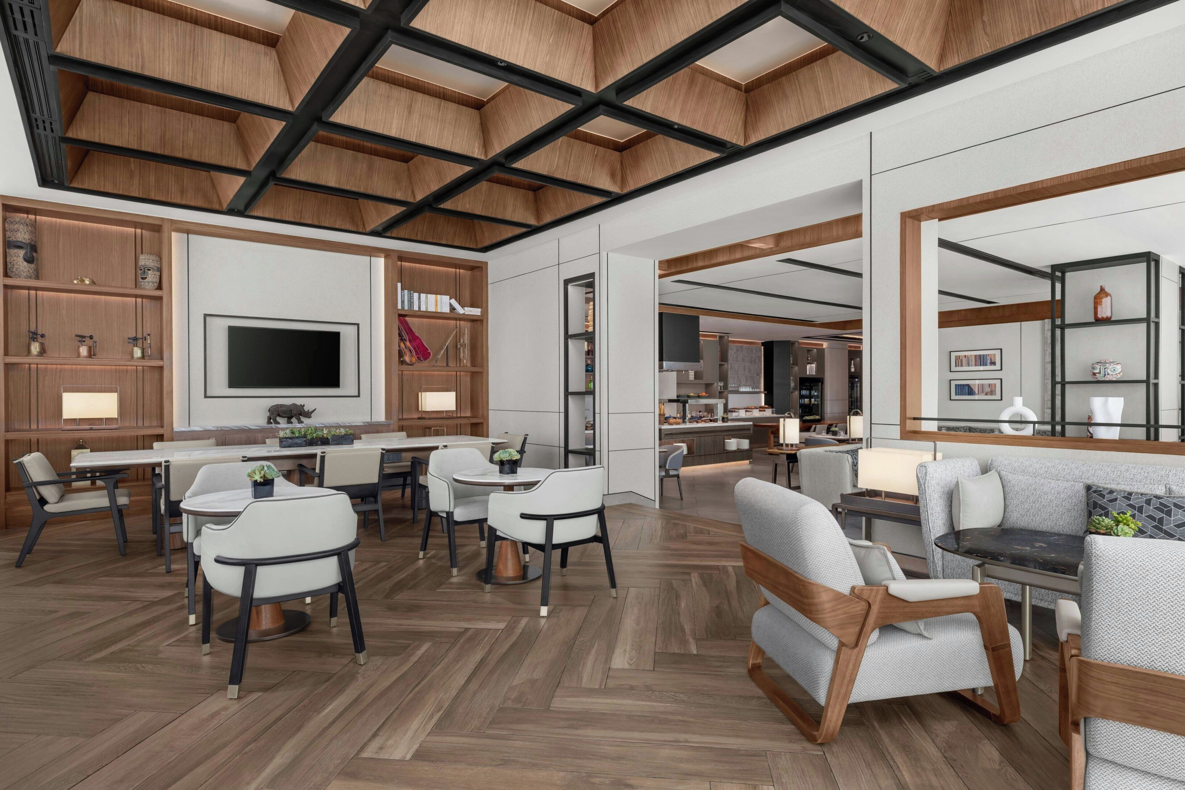 A modern interior with a herringbone floor, wooden ceiling beams, a central dining area with white chairs, and a kitchen area visible through an open doorway.