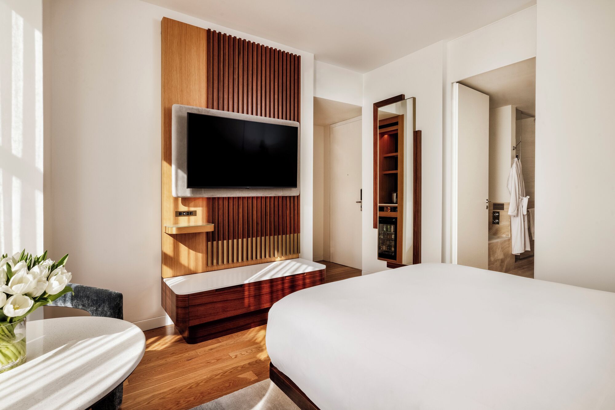 A modern hotel room with a large bed, a flat-screen TV mounted on a wooden panel, a small seating area with a table, and a bathroom visible through an open door.