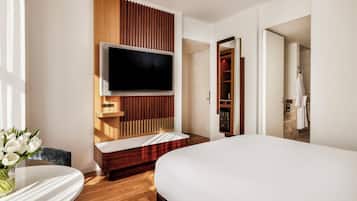 A modern hotel room with a large bed, a flat-screen TV mounted on a wooden panel, a small seating area with a table, and a bathroom visible through an open door.