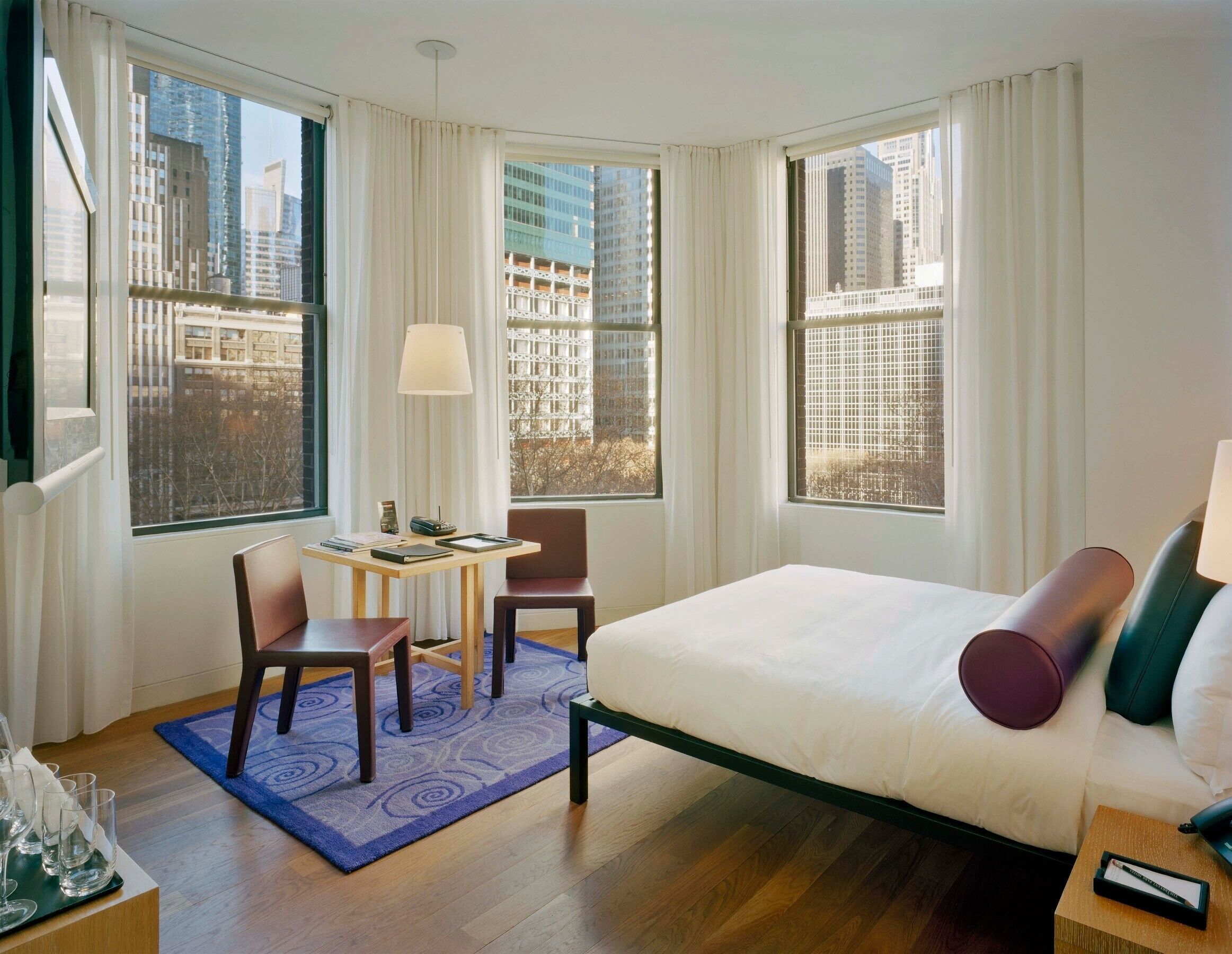A hotel room with a bed, two chairs, a small table, and a window with a city view.