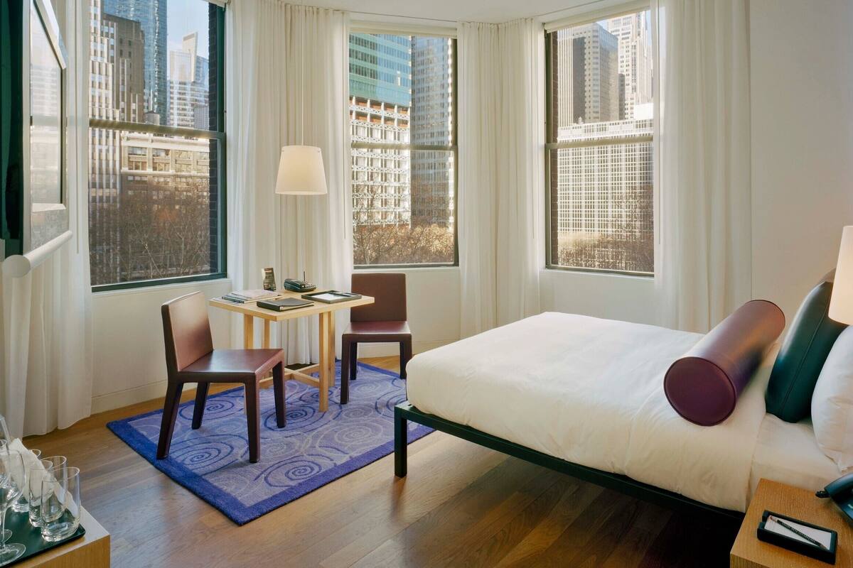 A hotel room with a bed, two chairs, a small table, and a window with a city view.