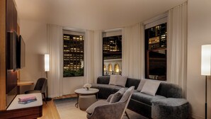 Una sala de estar moderna con un sofá gris, una mesa de centro redonda y una silla. Hay un televisor montado en la pared, una lámpara de pie y vistas de un paisaje urbano a través de amplias ventanas.