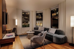Uma sala de estar moderna com um sofá cinza, uma mesa de centro redonda e uma cadeira. Há uma televisão na parede, um abajur e vista para um horizonte urbano através de grandes janelas.
