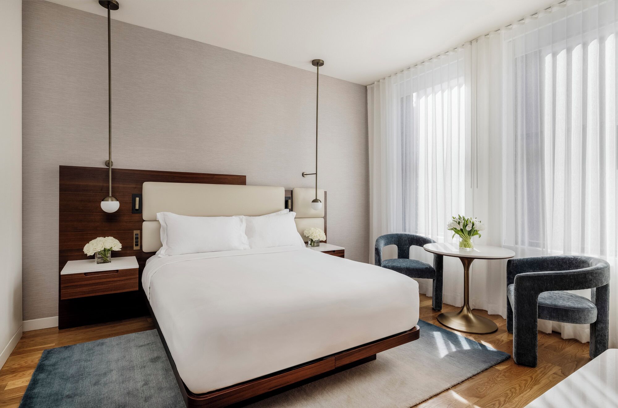 A modern hotel room with a large bed, bedside tables, a round table with chairs, and a view of the outdoors through sheer curtains.