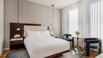 A modern hotel room with a large bed, bedside tables, a round table with chairs, and a view of the outdoors through sheer curtains.
