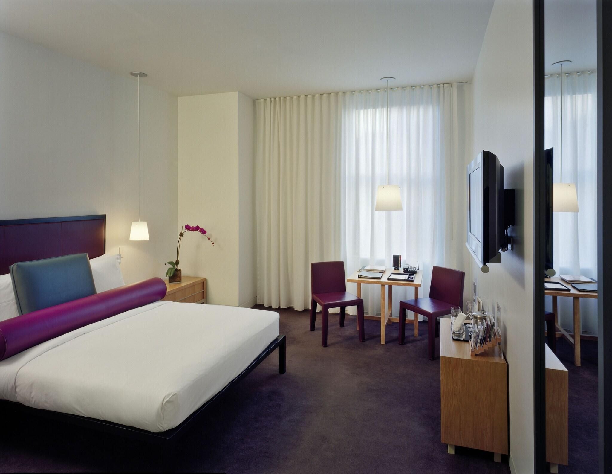 A hotel room with a bed, a desk with two chairs, a TV, and a window with curtains.