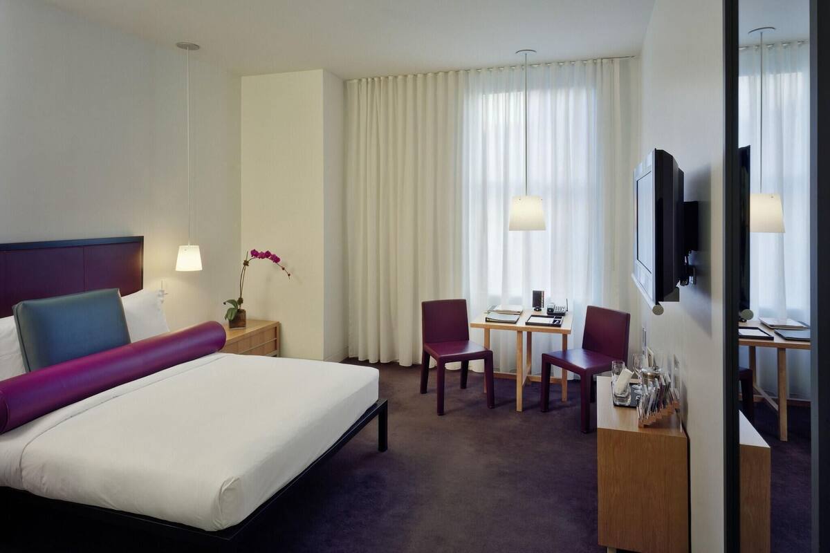 A hotel room with a bed, a desk with two chairs, a TV, and a window with curtains.