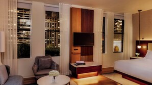 Habitación de hotel moderna con una cama grande, un sofá, una pequeña mesa redonda con un jarrón de flores, un televisor montado en un panel de madera y ventanas amplias con vistas a la ciudad.