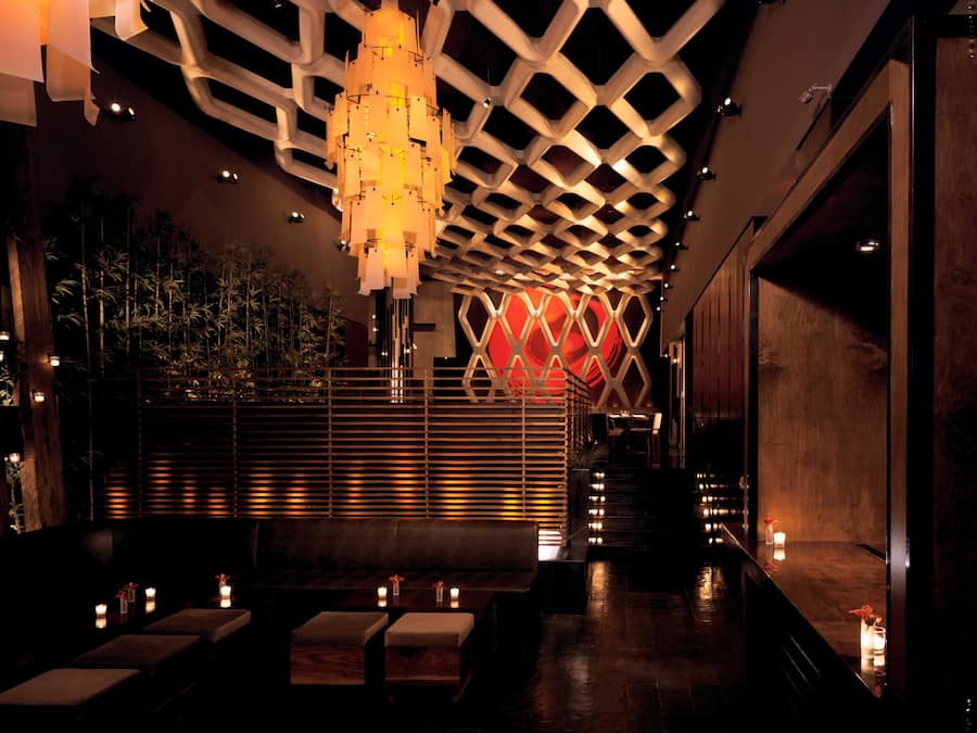 A dimly lit interior with a modern geometric ceiling design, a seating area with cushioned benches, and a wooden floor.