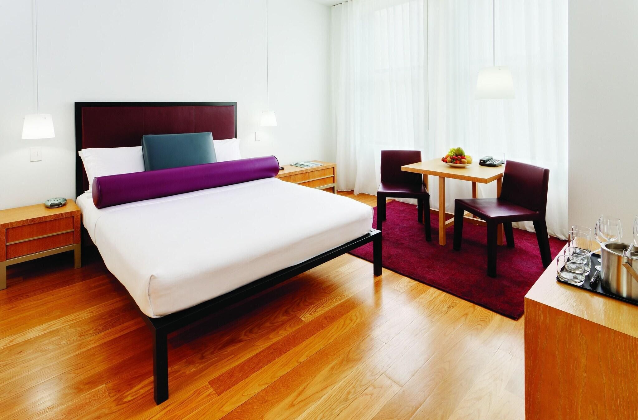 A hotel room with a bed, bedside tables, a desk with a chair, a small table with a fruit bowl, and a window with curtains.