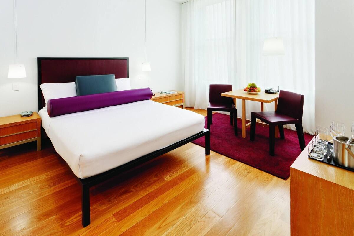 A hotel room with a bed, bedside tables, a desk with a chair, a small table with a fruit bowl, and a window with curtains.