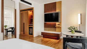 A modern hotel room with a large bed, a TV mounted on a wooden panel wall, a seating area with a table and chairs, and a minibar.