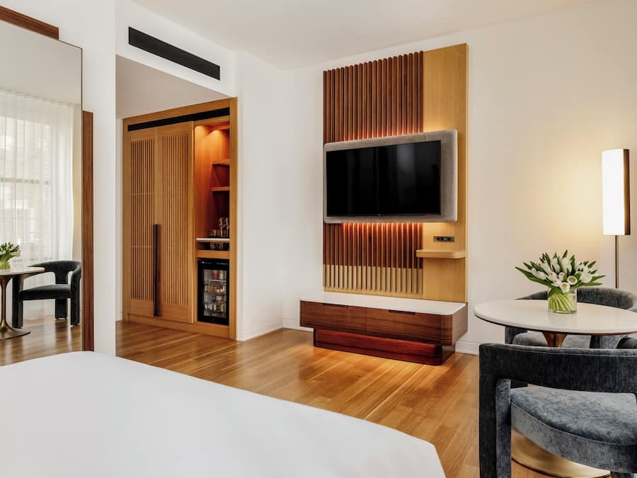 A modern hotel room with a large bed, a TV mounted on a wooden panel wall, a seating area with a table and chairs, and a minibar.