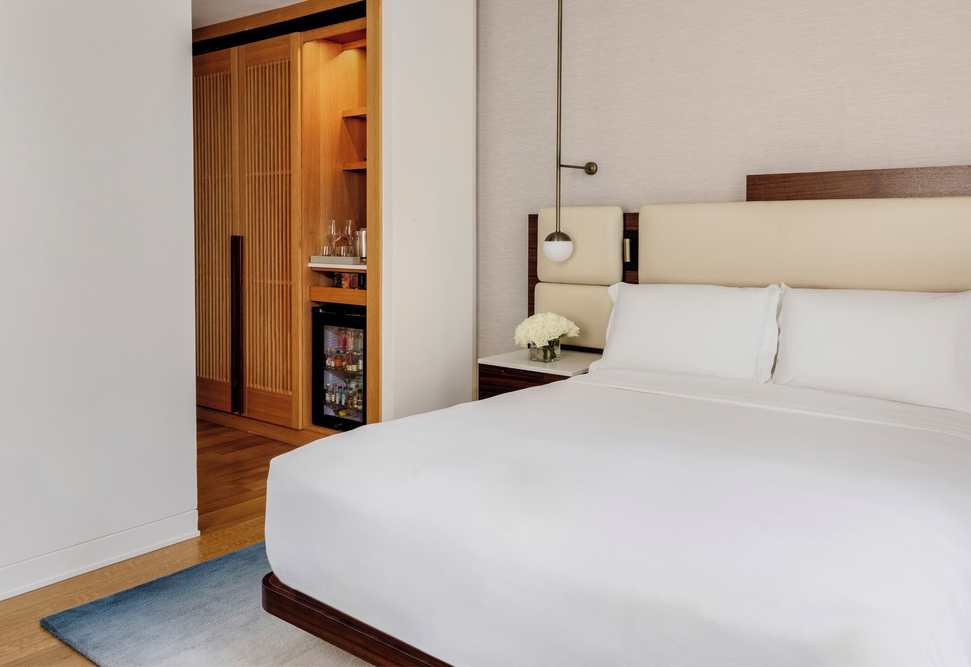 A hotel room with a large bed, a bedside table with a lamp, a wooden wardrobe, and a small shelf with items.