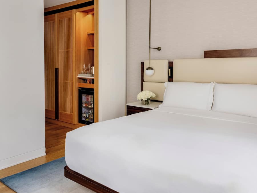 A hotel room with a large bed, a bedside table with a lamp, a wooden wardrobe, and a small shelf with items.